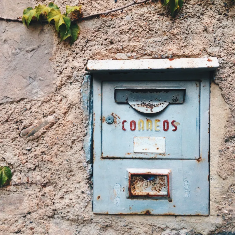 A weathered blue Correos mailbox set into a stone wall with ivy, from Lucienne's travels.