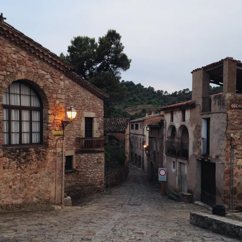 A cobblestone street between old stone houses in a village in Spain, with a lit street lamp at dusk.